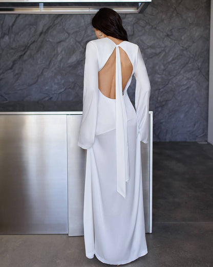 Woman wearing a white open-back dress standing against a dark textured wall.