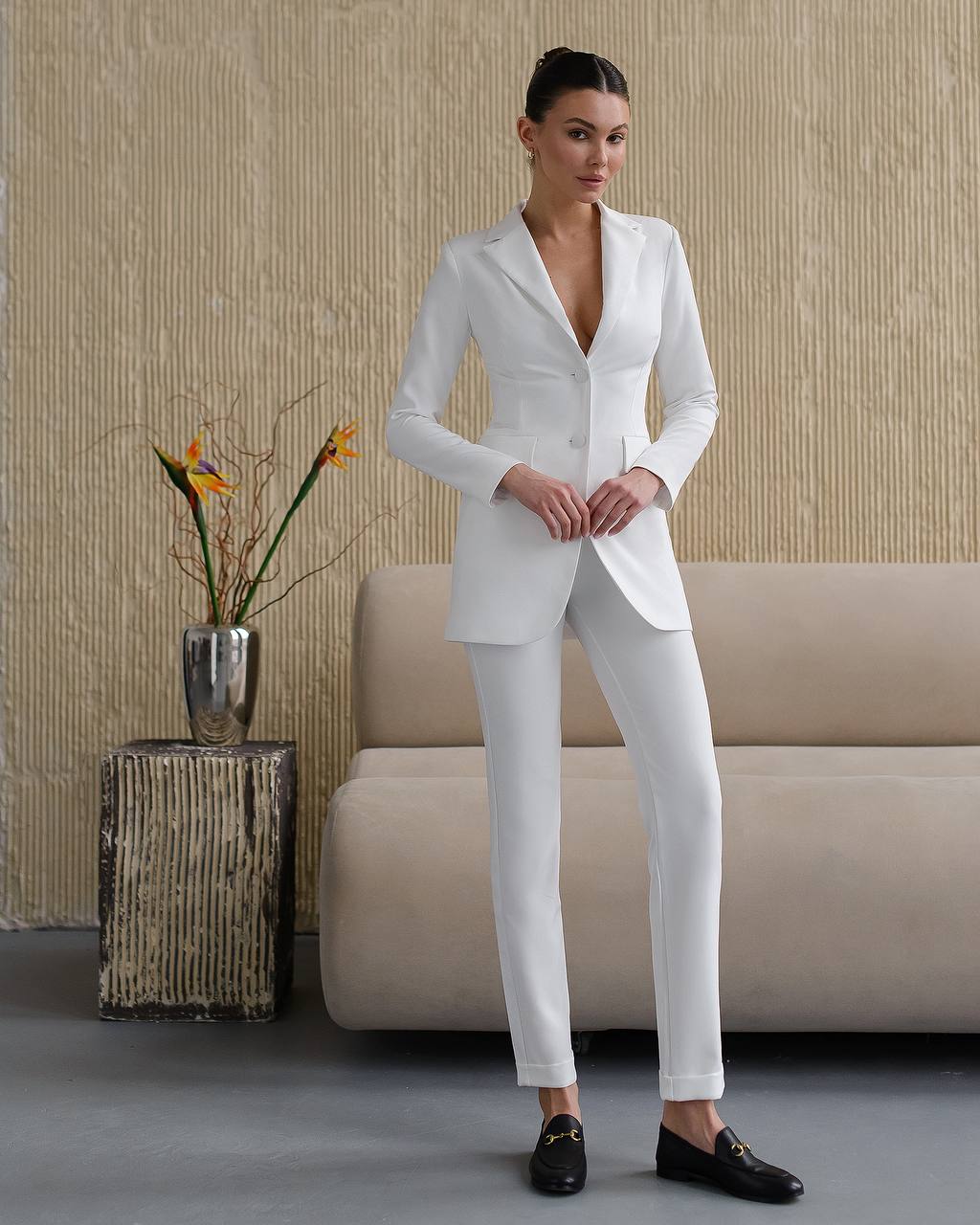 Woman wearing a white suit standing in a room with a beige sofa and decorative plant.