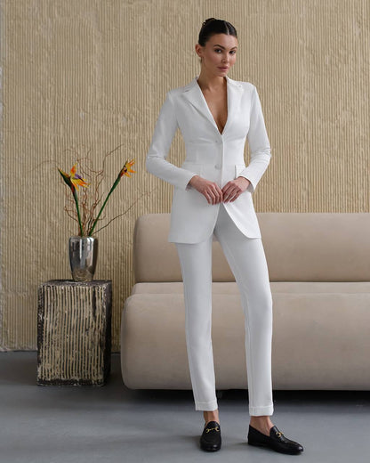 Woman wearing a white suit standing in a room with a beige sofa and decorative plant.