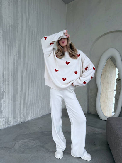 Person wearing a white sweatshirt with red heart patterns and white pants in a minimalistic room.