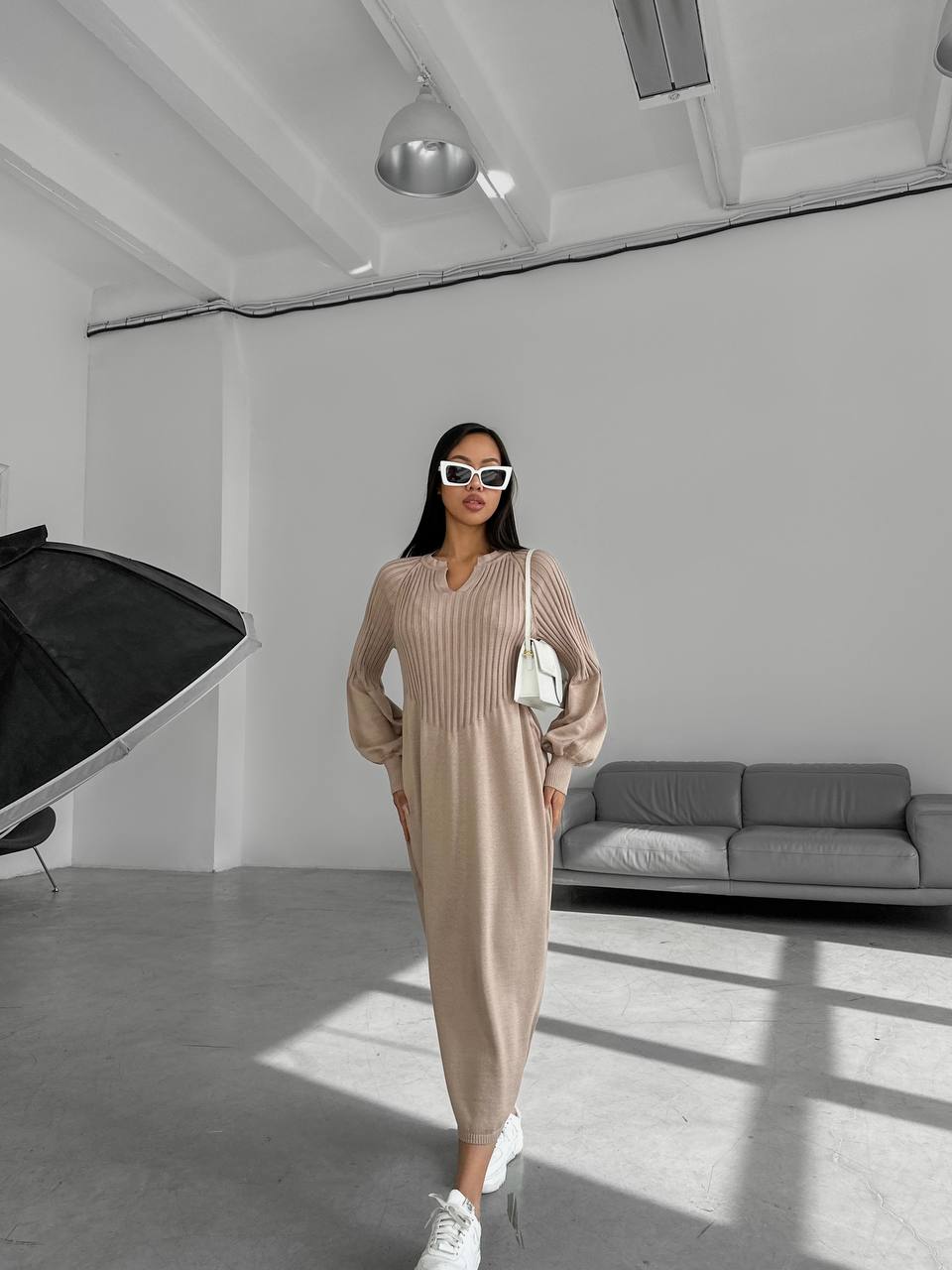 Woman in a beige outfit standing in a modern room with a gray sofa and black chair.