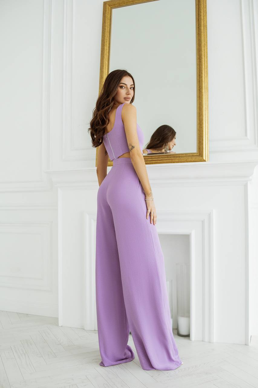 Woman in a lavender jumpsuit standing in front of a mirror on a white wall.