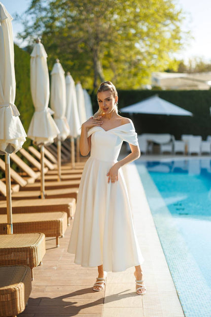 Woman in a white dress standing by a poolside with umbrellas and lounge chairs in the background.