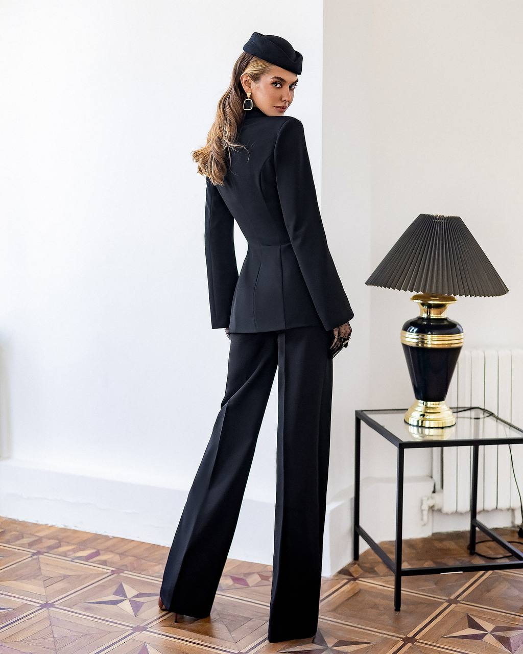 Woman in a black suit standing in a room with a lamp on a table.