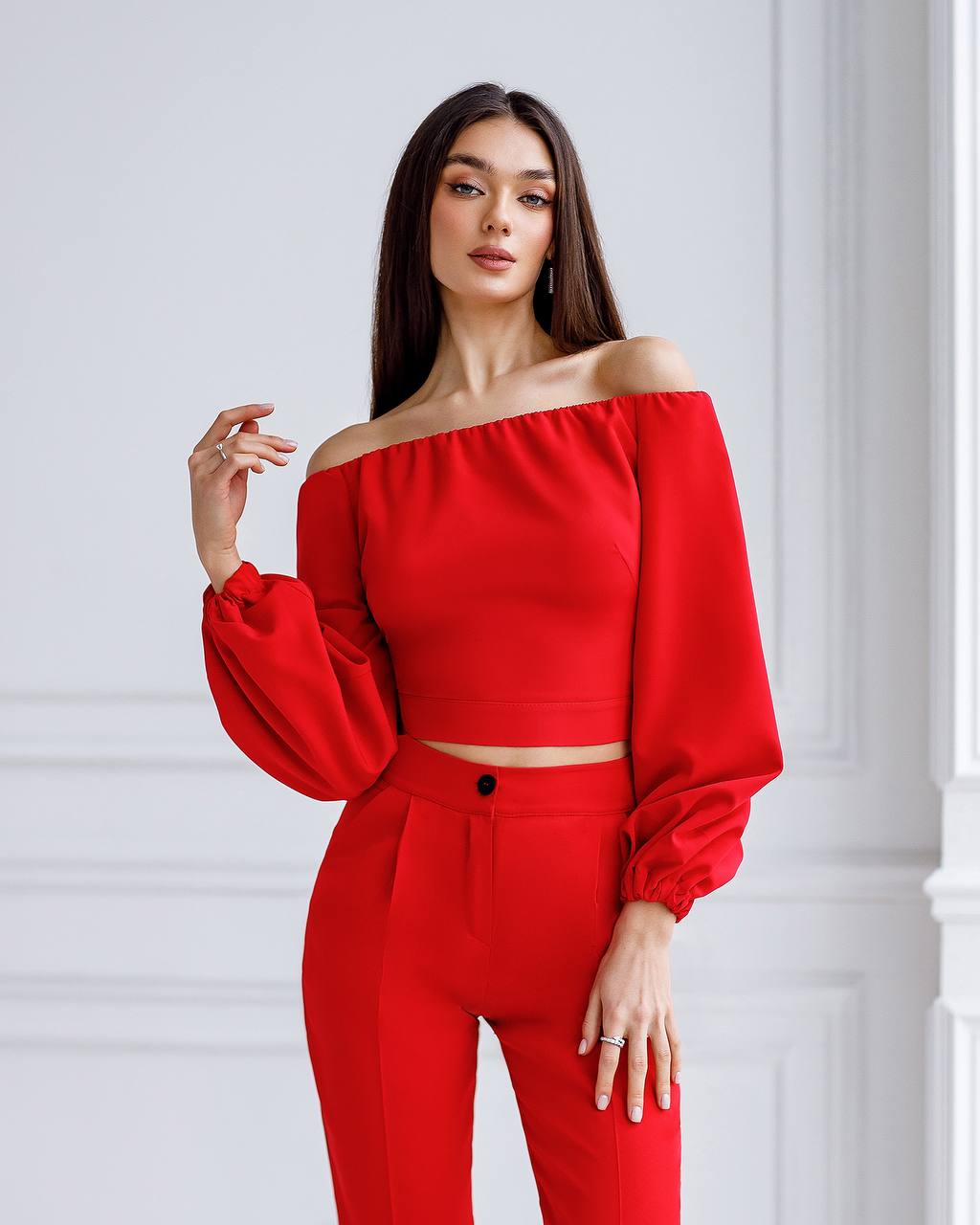 Woman wearing a red outfit with off-shoulder top and high-waisted pants against a white wall.