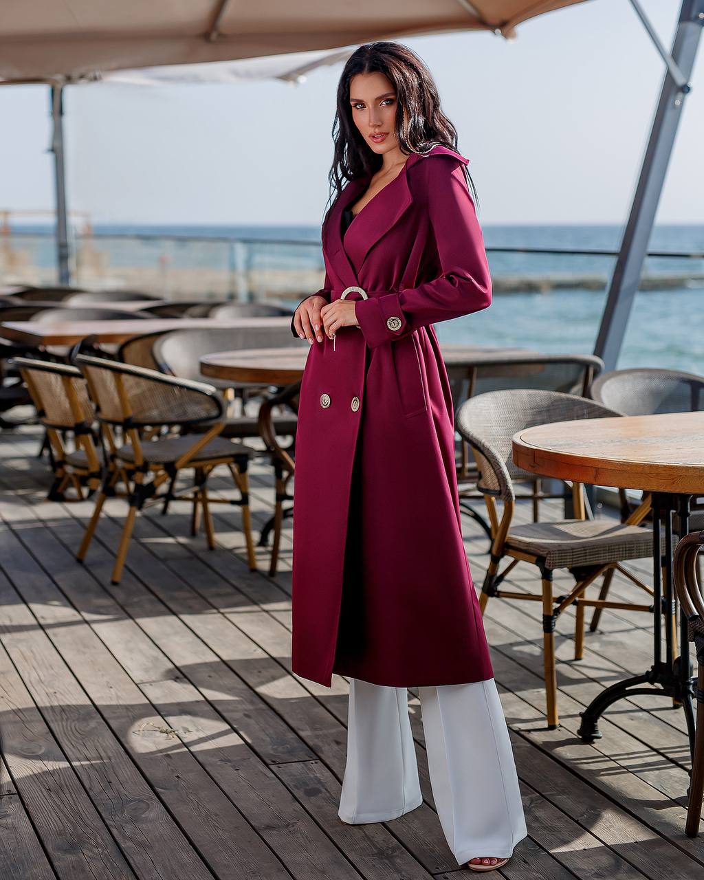 Woman in a long burgundy coat standing on a wooden deck with tables and chairs, near water.
