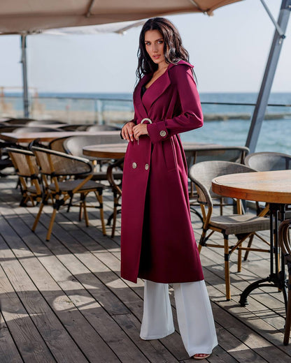 Woman in a long burgundy coat standing on a wooden deck with tables and chairs, near water.