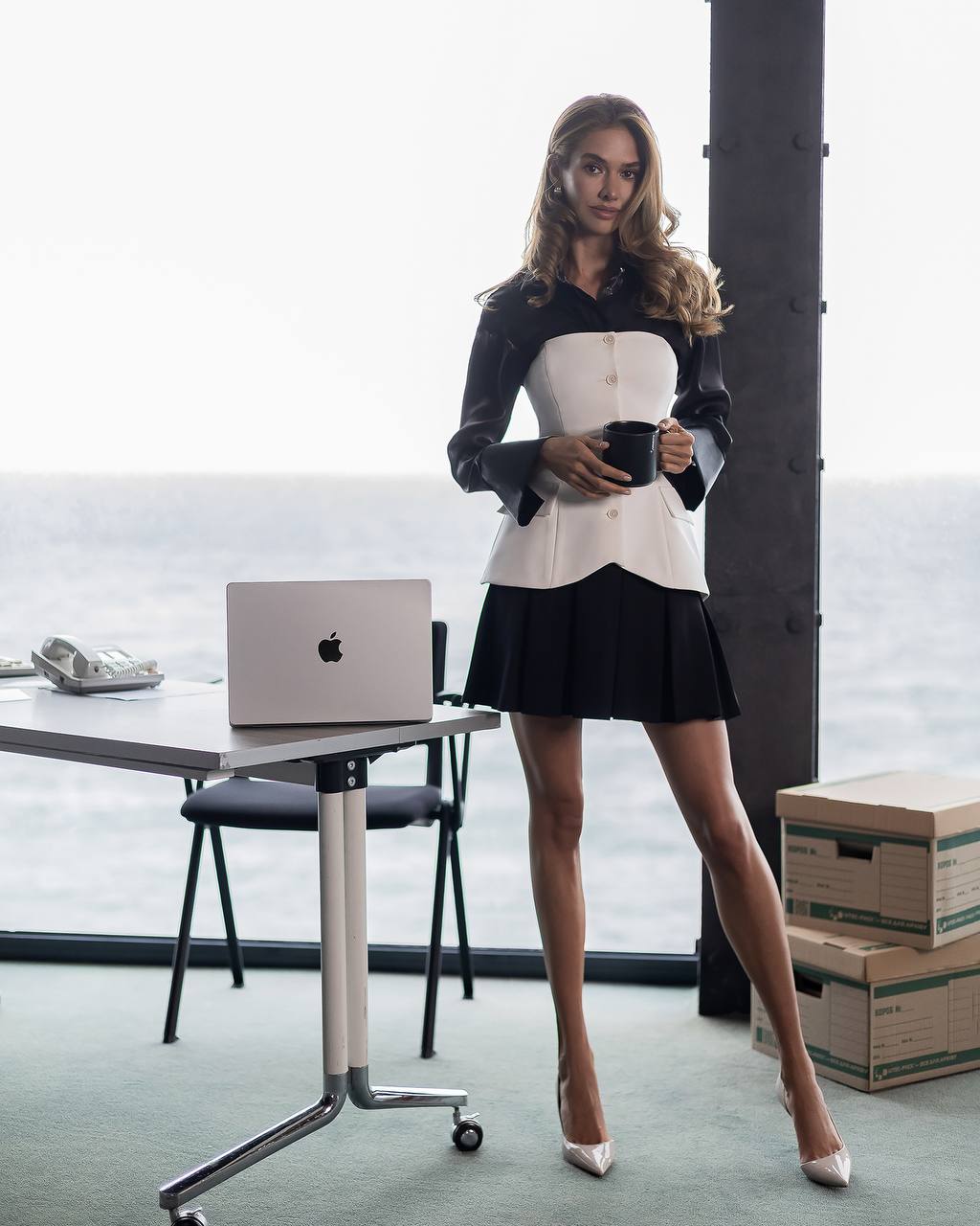 A woman stands in a bright office-like space by a large window, wearing a black and white mini dress with structured shoulders and holding a dark mug. A silver laptop sits on a table beside her, with cardboard boxes stacked near the window.