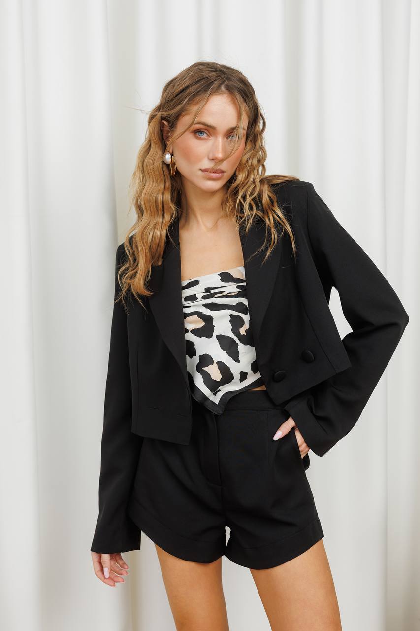 Woman wearing a black blazer, leopard print top, and black shorts against a white curtain background