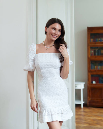 Woman wearing a white dress standing indoors with a wooden bookshelf in the background