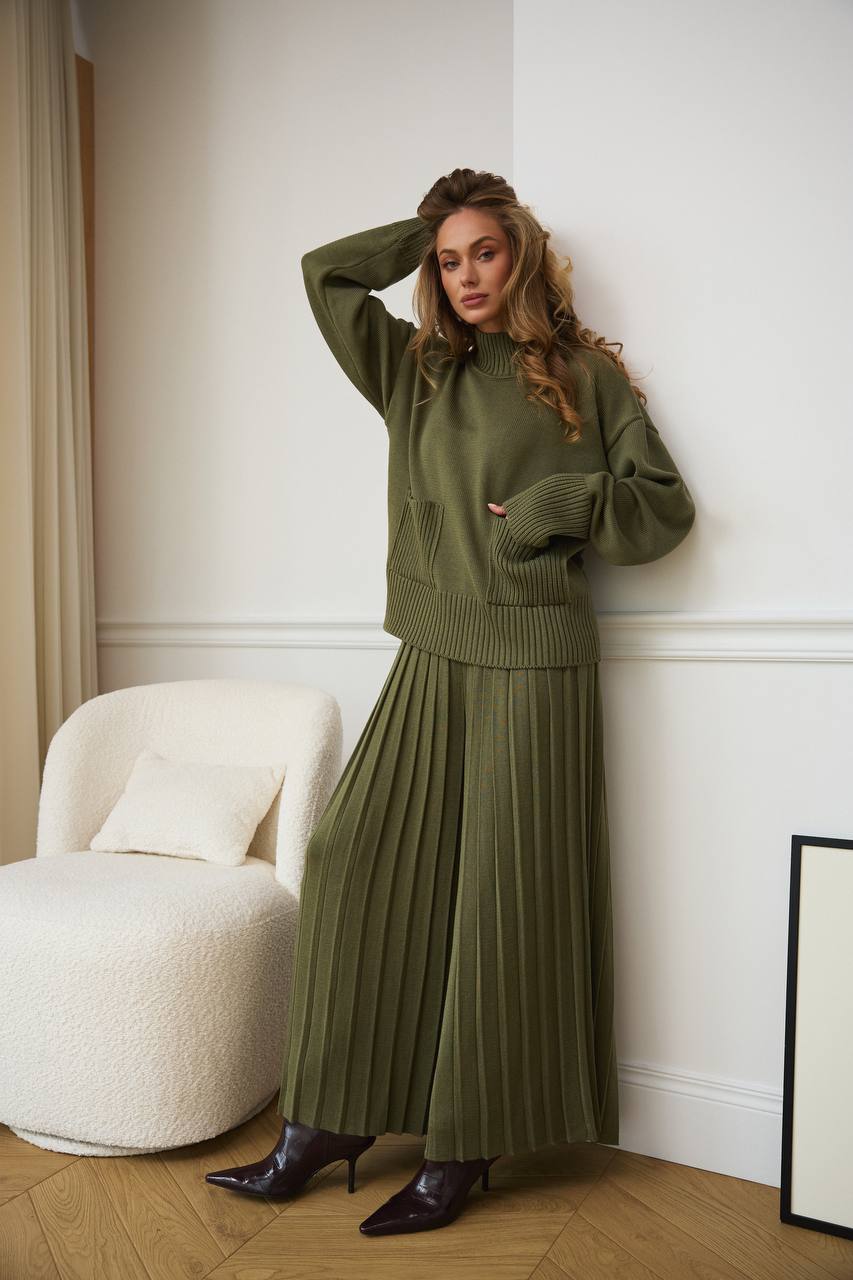 Woman wearing a green knit outfit standing in a room with a white chair and light-colored walls.