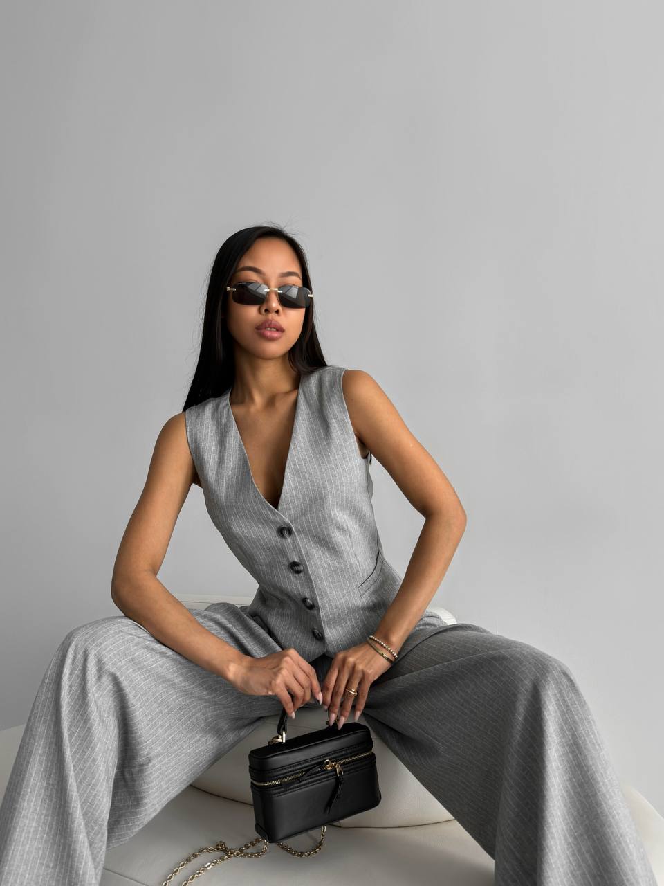 Woman in a gray sleeveless top and wide-leg pants holding a black handbag against a gray background