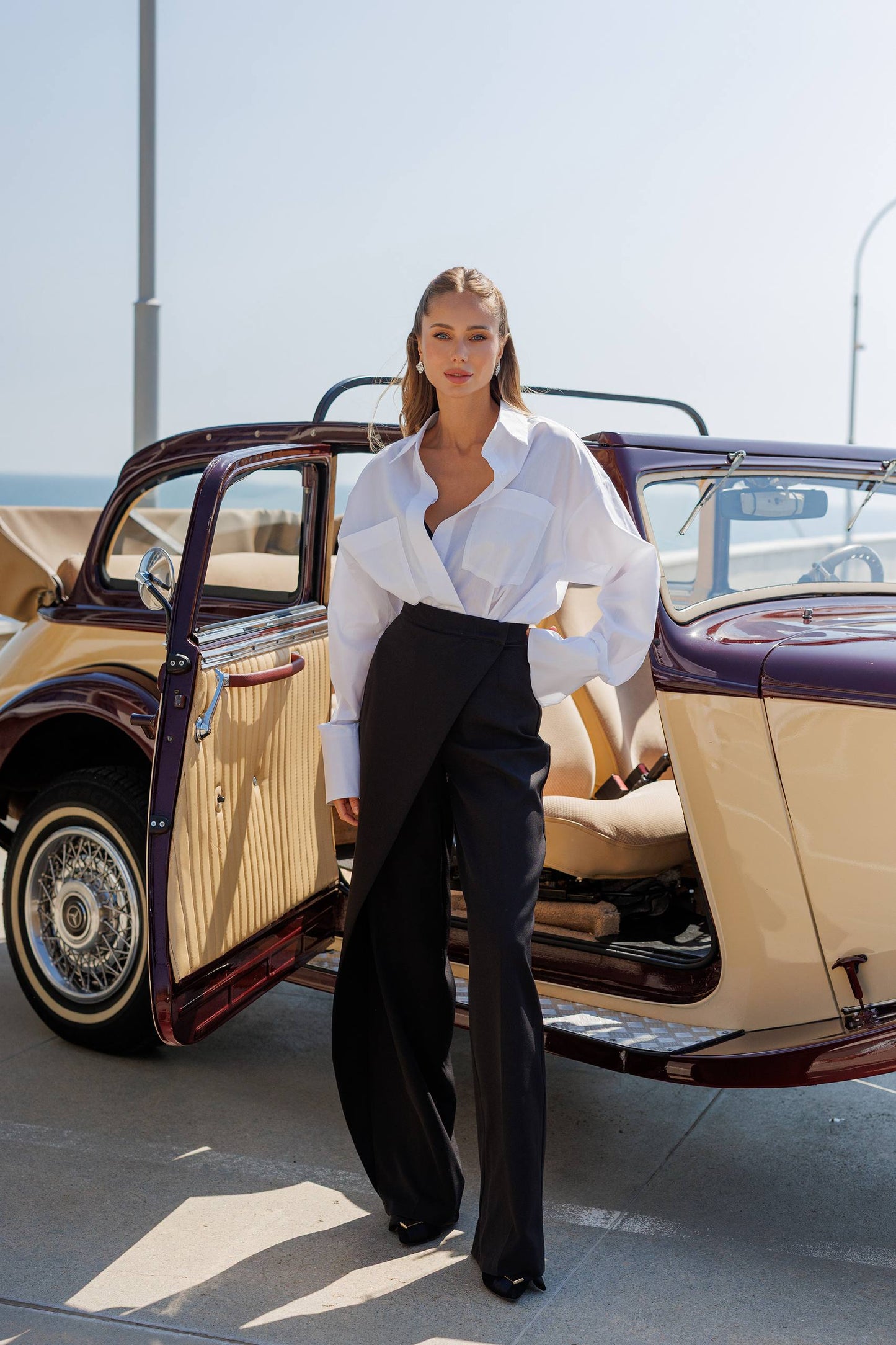 Woman in a white blouse and black pants standing next to a vintage car.