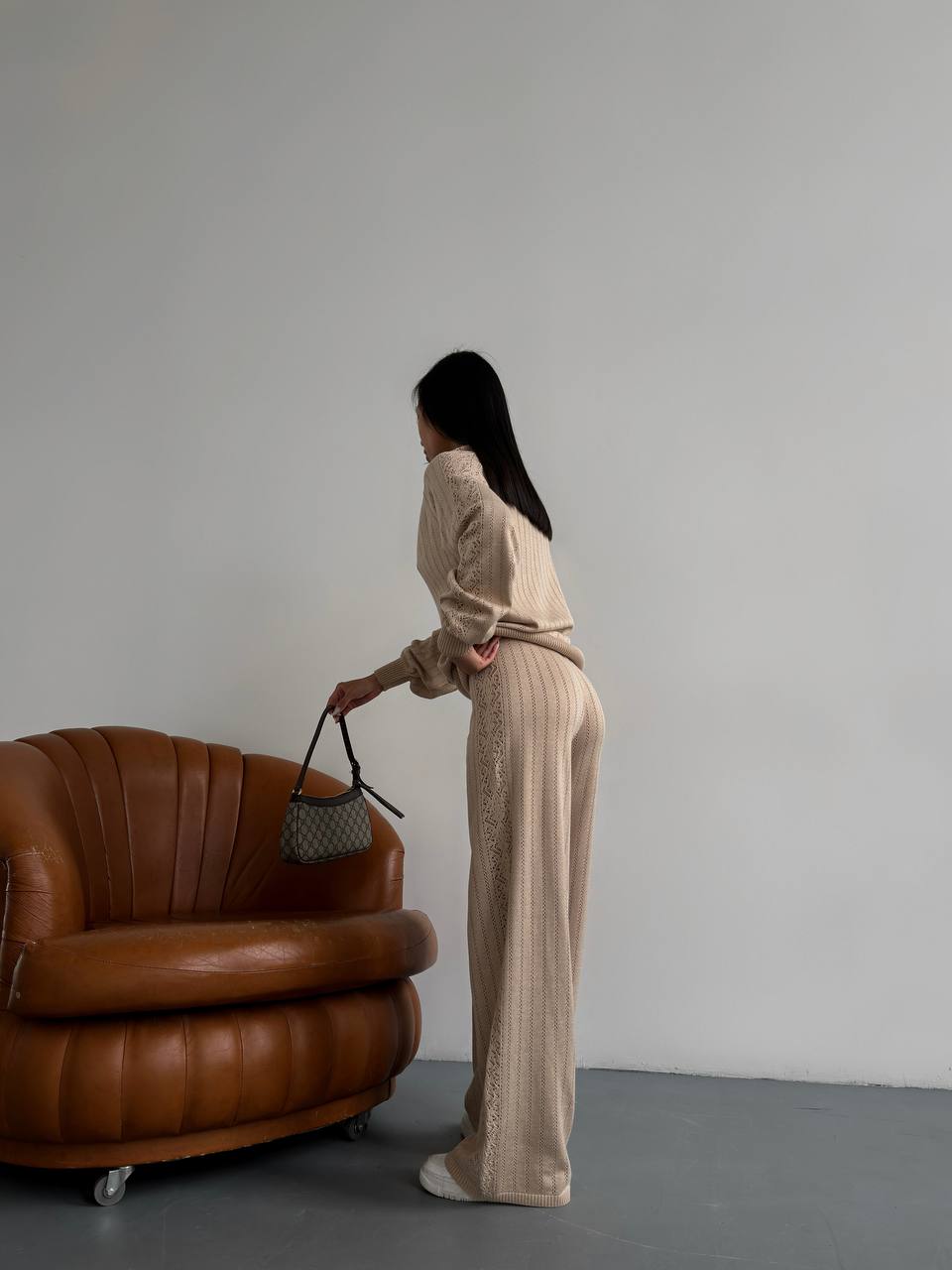 Woman in a beige outfit standing next to a brown leather sofa against a plain wall.