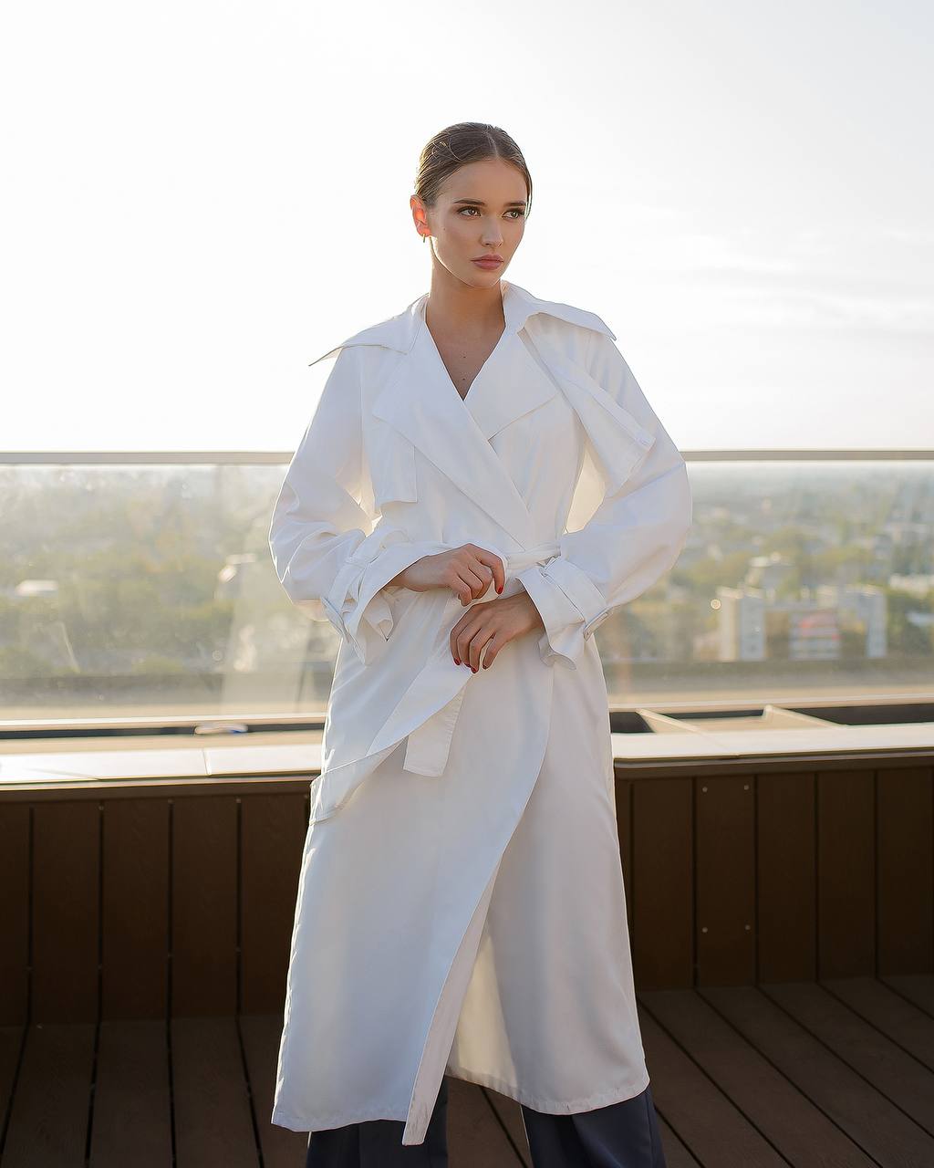 Woman wearing a white trench coat on a rooftop with a cityscape background