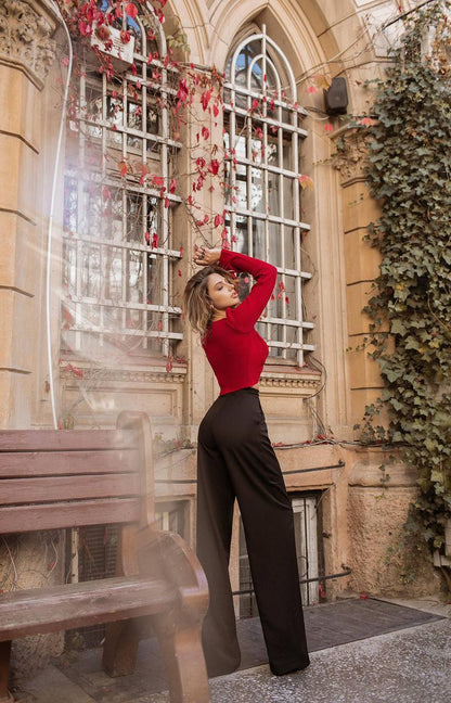 Woman in a red top and black pants posing in front of a decorative window with floral elements.