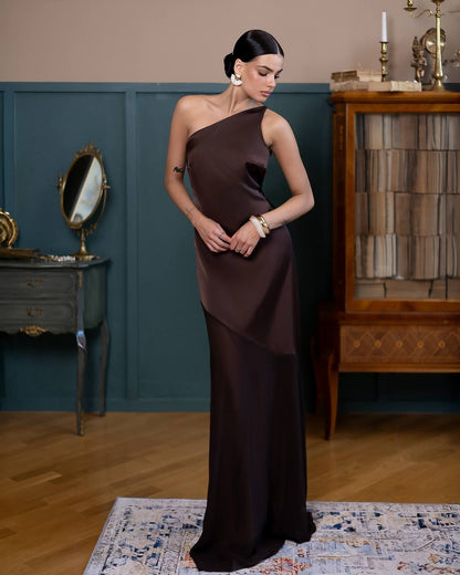 A woman models a floor-length, one-shoulder satin gown in a dark chocolate brown color, standing in a vintage-style room with teal wainscoting, a wooden cabinet, and a patterned rug.
