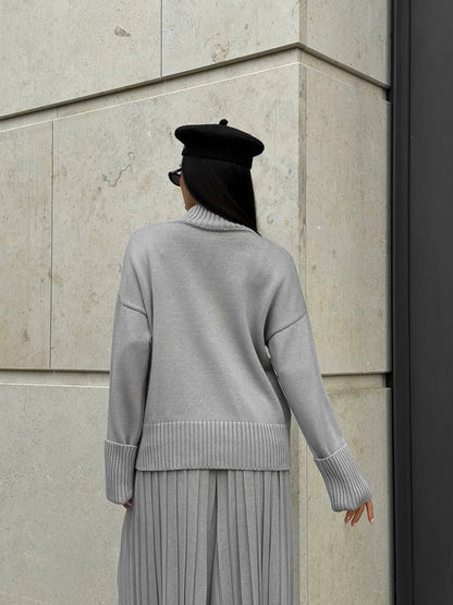 Person wearing a gray sweater and black beret against a stone wall.