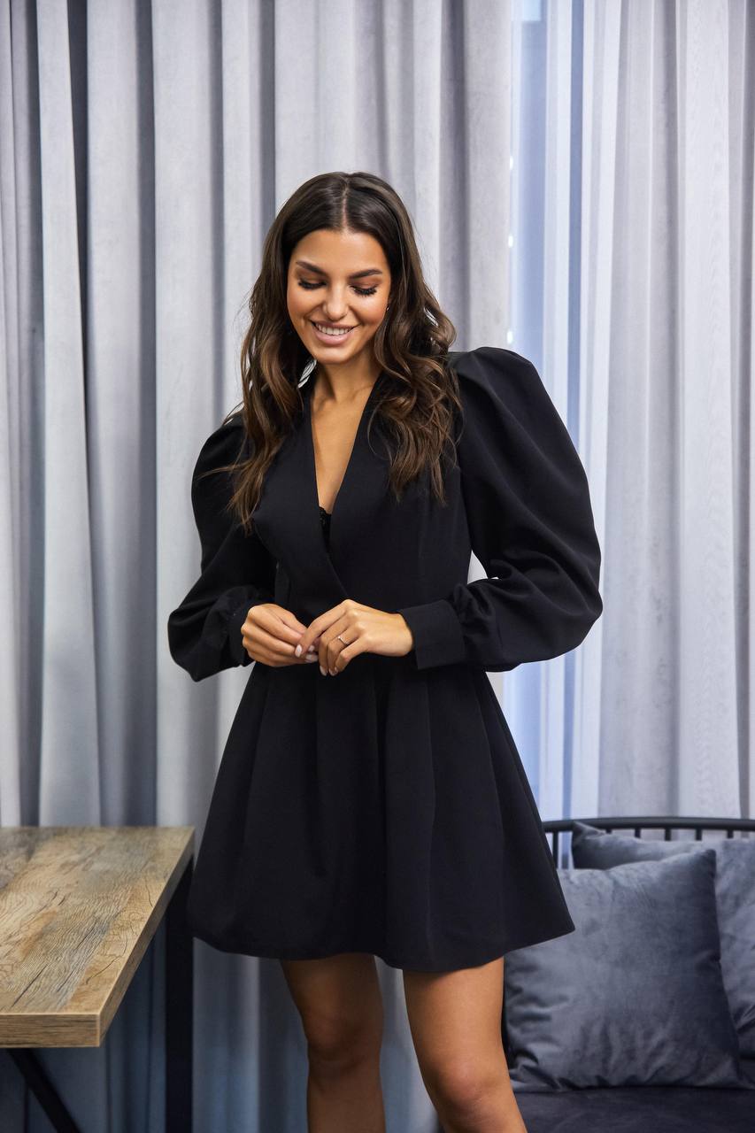 Woman wearing a black dress standing in a room with curtains and a couch.