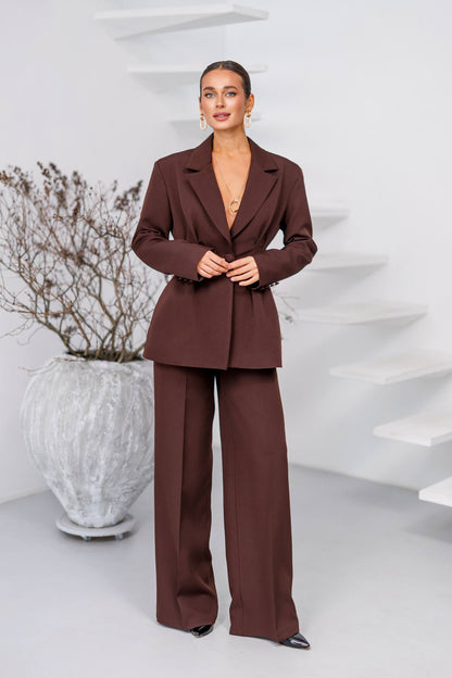 Woman wearing a brown suit standing in a minimalistic room with a white vase and plant.