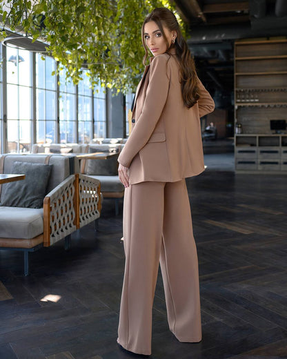Woman in a beige suit standing in a modern indoor setting with large windows and a tree.