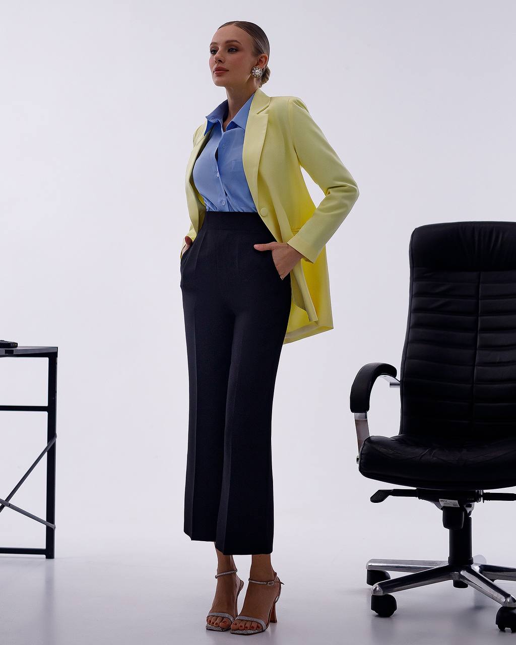A woman models a light yellow blazer over a blue button-down shirt and black wide-leg cropped trousers, standing in a studio next to a black office chair.