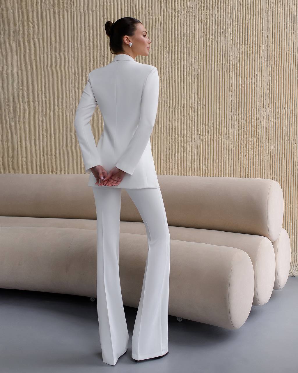 Woman in a white outfit standing in a modern room with beige sofa and wall.