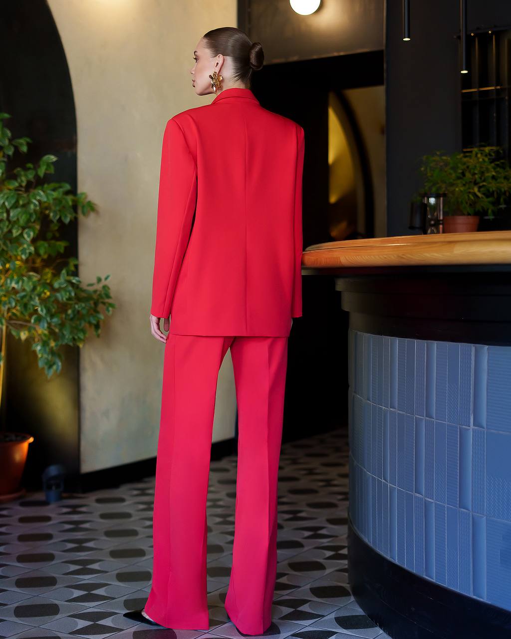 Woman in a red suit standing in a modern interior setting with plants and a counter.