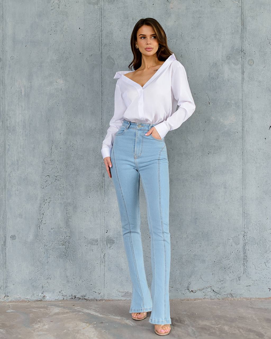 Woman wearing a white blouse and light blue jeans against a gray concrete wall.