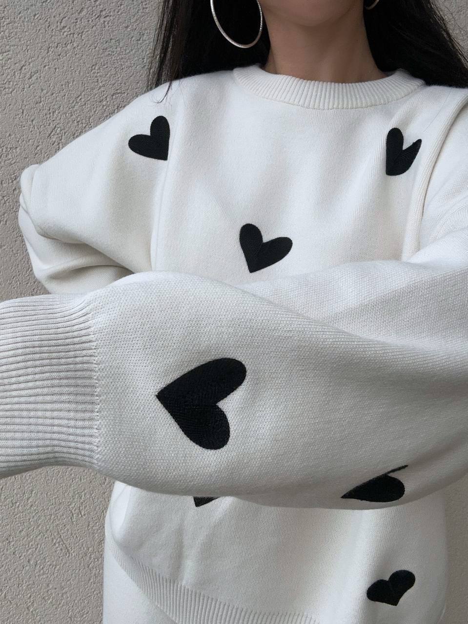 White sweater with black heart patterns worn by a person against a neutral background