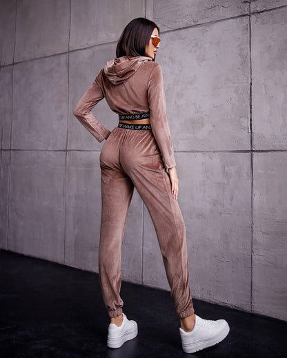 Woman wearing a brown tracksuit standing against a gray wall.