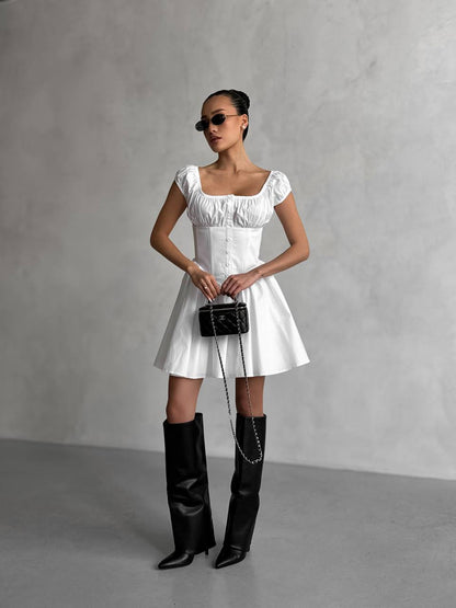 Woman in a white dress and black knee-high boots holding a black handbag against a gray background
