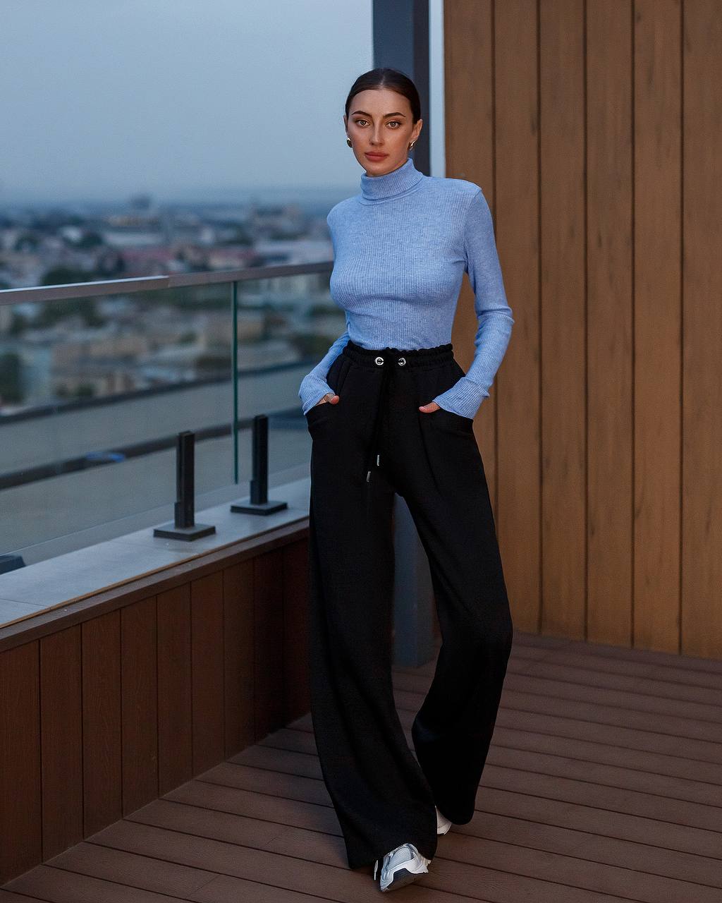 Woman in a light blue turtleneck and black pants standing on a balcony with a cityscape view.