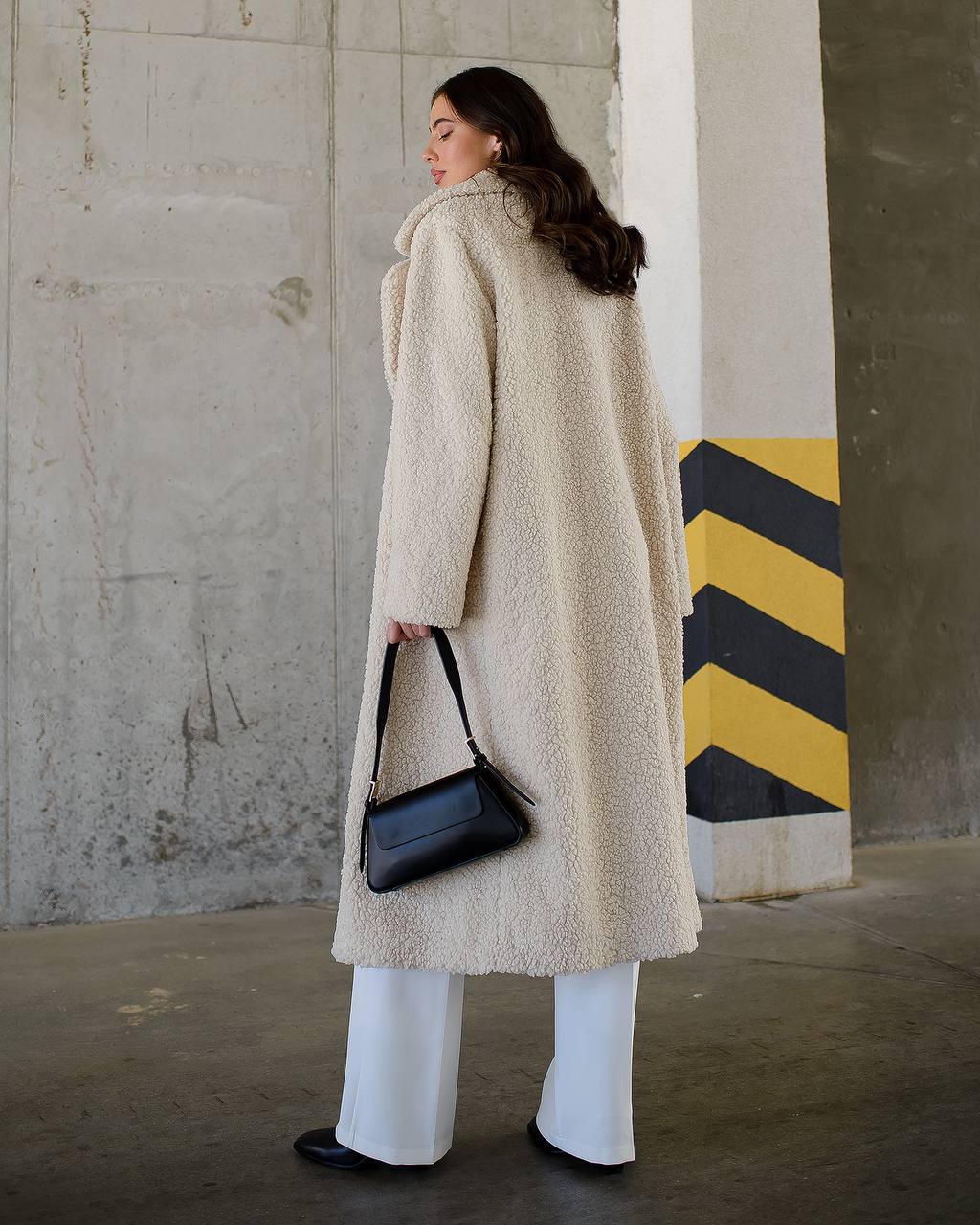 Woman wearing a long beige coat holding a black handbag against a concrete wall.