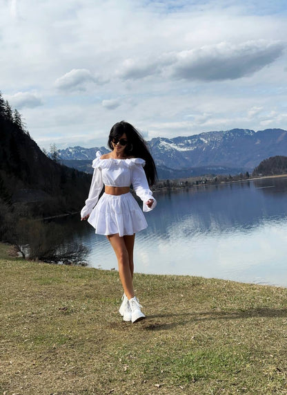 Woman in a white outfit standing by a lake with mountains in the background