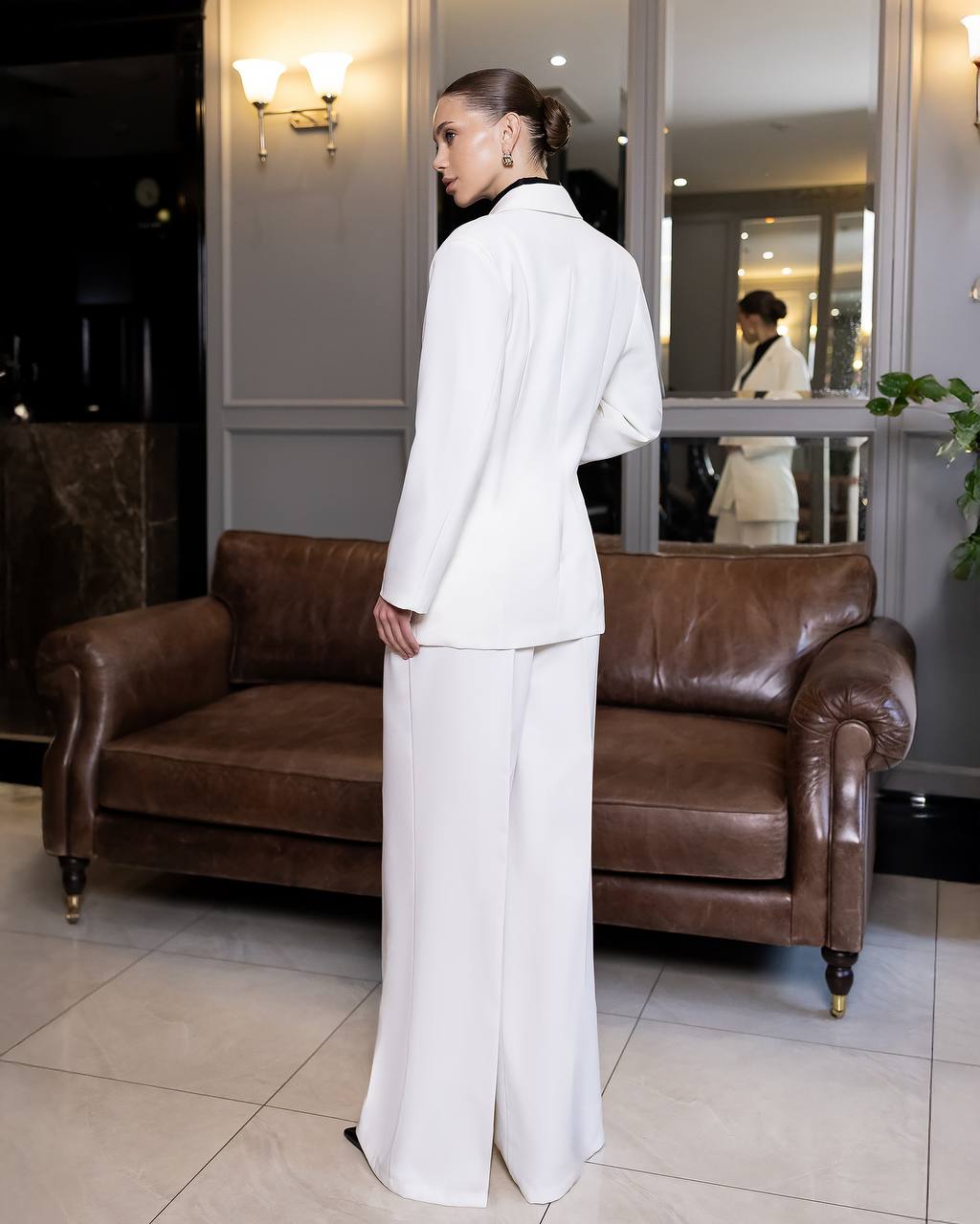 Woman in a white suit standing in a room with a brown leather sofa and mirror.