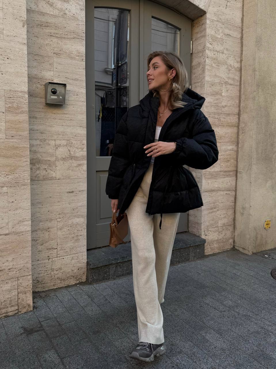 Woman in a black jacket and beige pants standing outside a building.