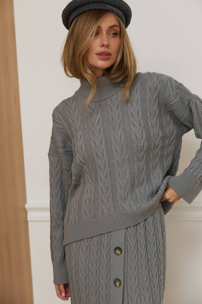 Woman wearing a gray cable knit sweater with buttons, standing against a neutral background.