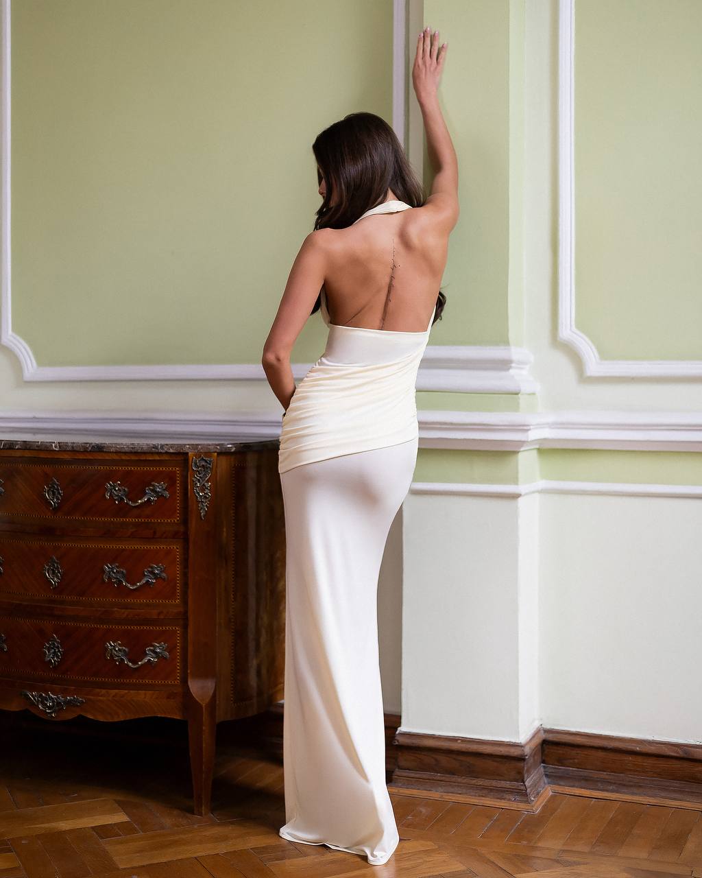 Woman in a white evening gown standing in a room with green walls and wooden furniture.