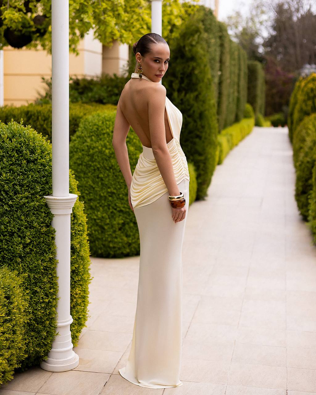 A woman wearing a champagne-colored, backless ruched gown stands on a garden pathway between trimmed green hedges.