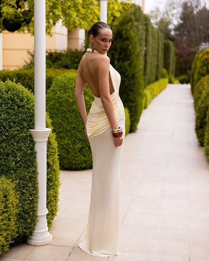 A woman wearing a champagne-colored, backless ruched gown stands on a garden pathway between trimmed green hedges.