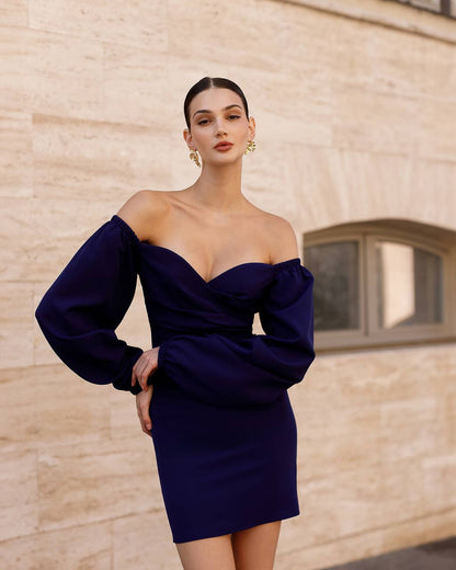 Woman wearing a dark blue off-shoulder dress against a beige brick wall.