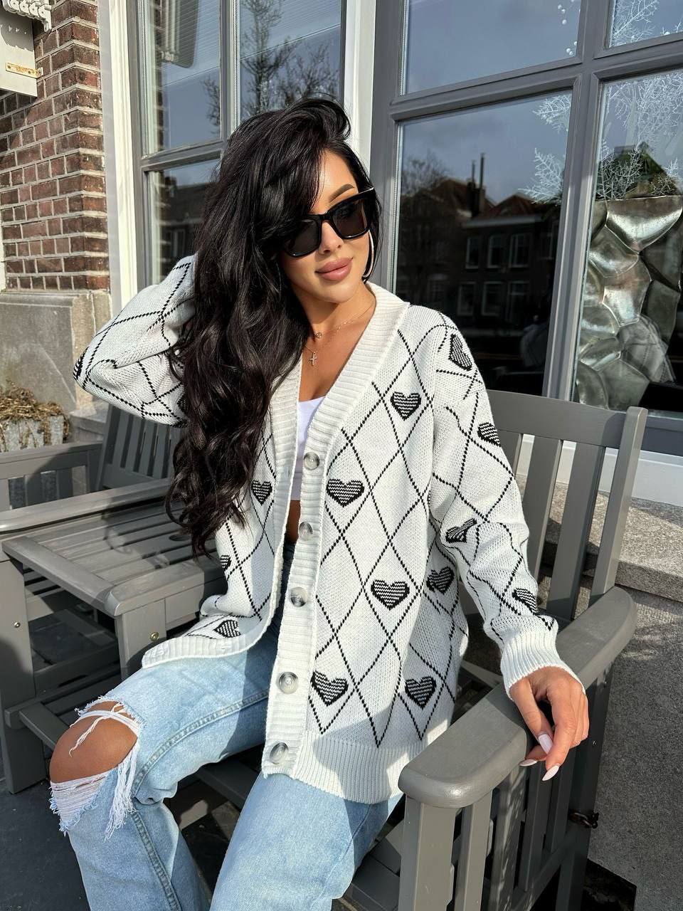 Woman wearing a white cardigan with black heart patterns, sitting outdoors.