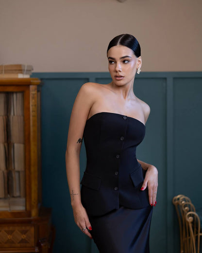 A woman modeling a strapless black corset-style dress with front buttons and a fitted silhouette, standing indoors against a dark teal paneled wall with a wooden cabinet on the left.