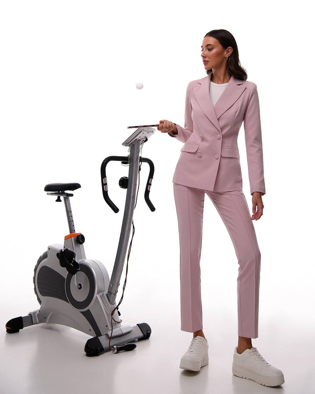 Woman in a pink suit standing next to an exercise bike on a white background