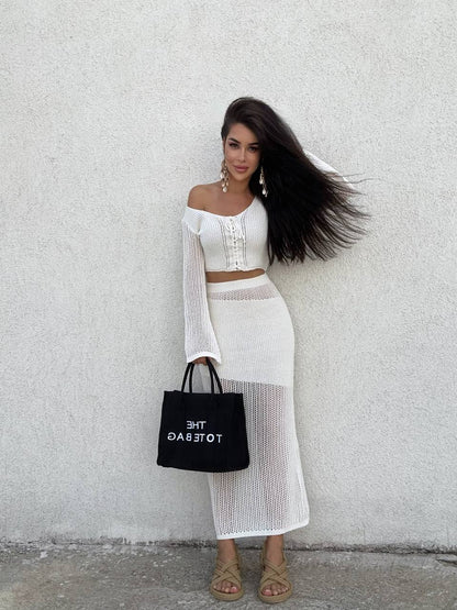 Woman in a white outfit holding a black tote bag against a light gray wall.