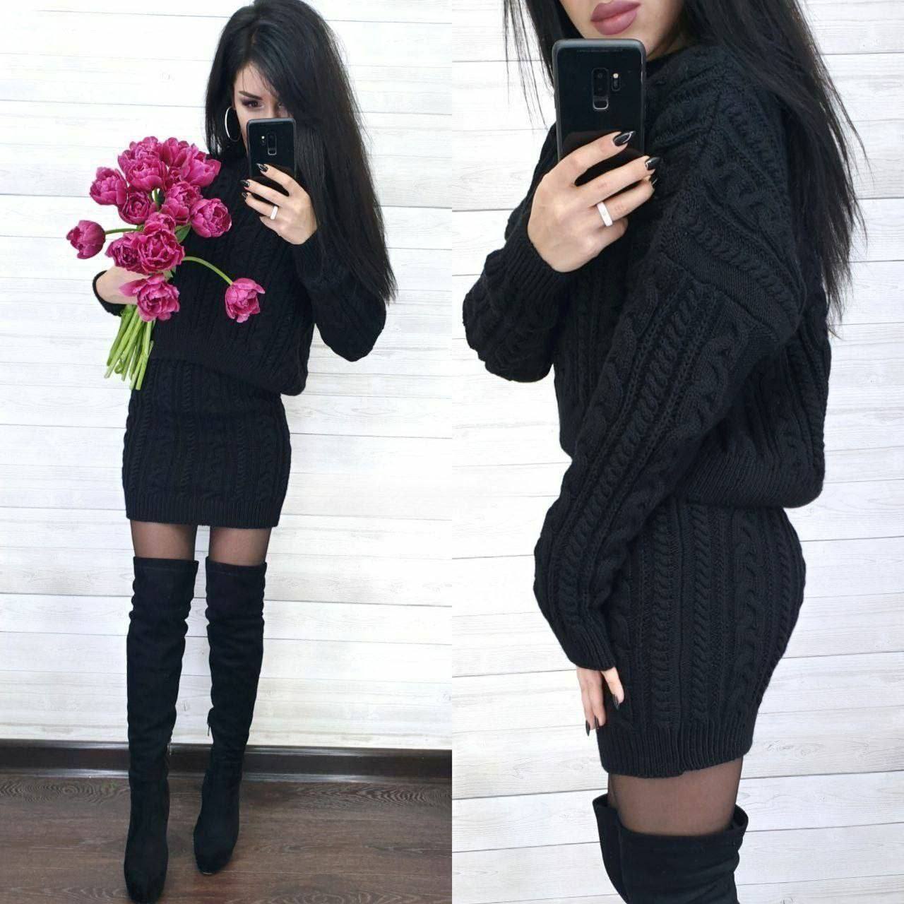 Woman wearing a black cable knit sweater dress with knee-high black boots, holding pink flowers.