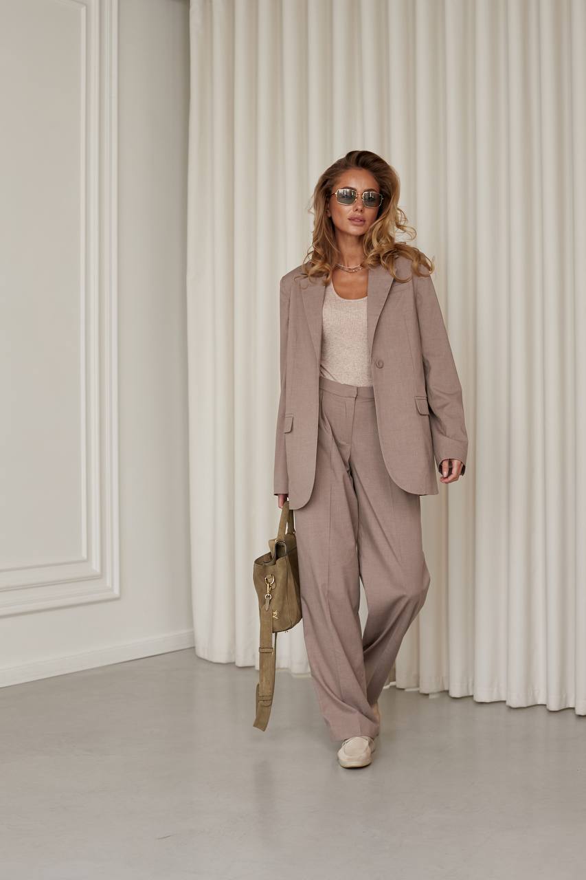 Woman in a beige suit standing in a room with white walls and curtains.
