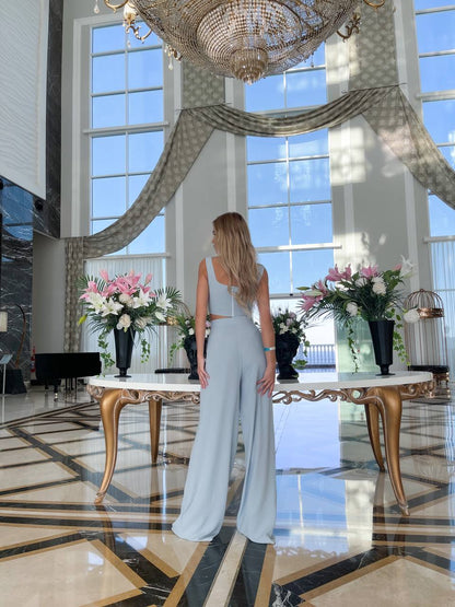 Woman in a light blue dress standing in a luxurious room with large windows and decorative flowers.