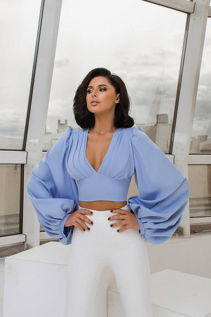 Woman wearing a light blue top with wide sleeves and white pants in an indoor setting.